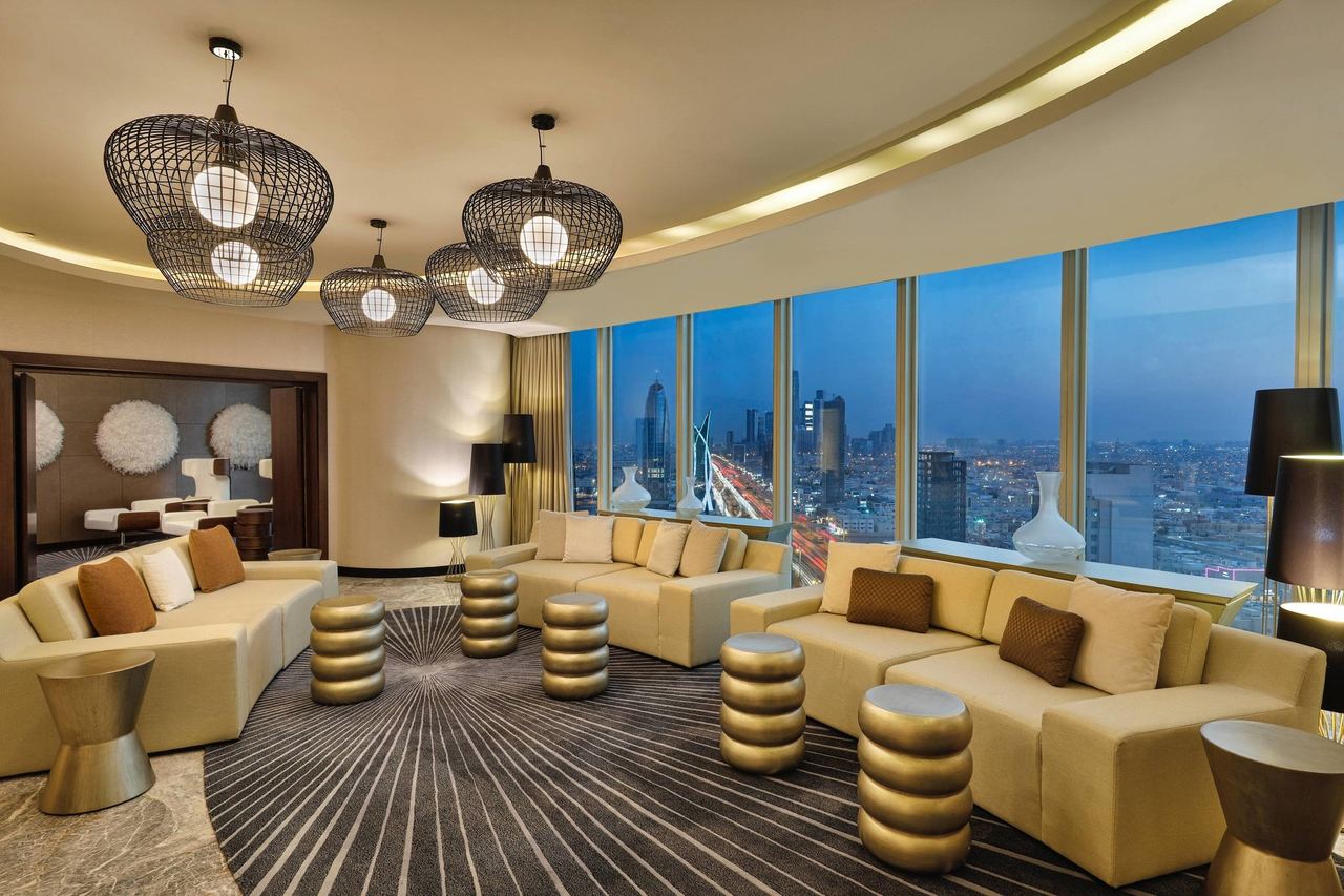 A suite lounge at JW Marriott Hotel Riyadh, curved sofas below wicker lanterns and a wall of glass over King Fahad Road at dusk