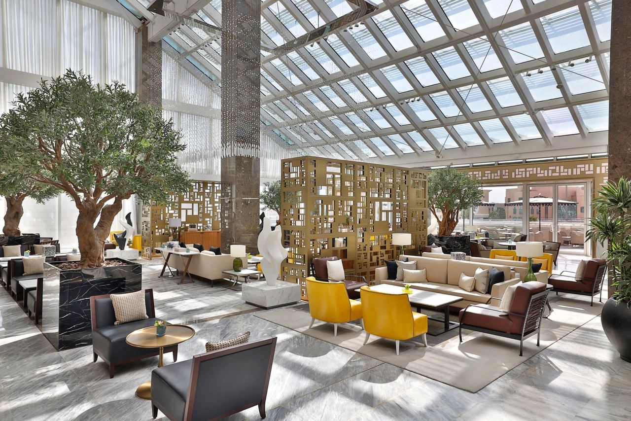 The lobby lounge of Sofitel Riyadh under its glass roof, olive trees and yellow armchairs on pale marble